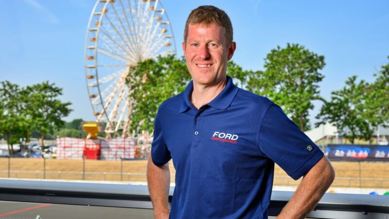 A man in a blue Ford Performance polo stands smiling outdoors with a Ferris wheel, trees, and a clear sky in the background, capturing the excitement of Ford Le Mans.
