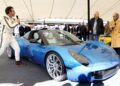 A man in a racing suit speaks into a microphone next to a blue sports car under a tent at the Festival of Speed, with an audience and two men standing nearby.