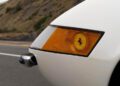 Close-up of the front right corner of a white 365 GTB/4 Daytona, showing an amber indicator light with a Ferrari logo and part of a chrome bumper. A road and hillside are in the background, evoking classic Ferrari Beverly Hills elegance.
