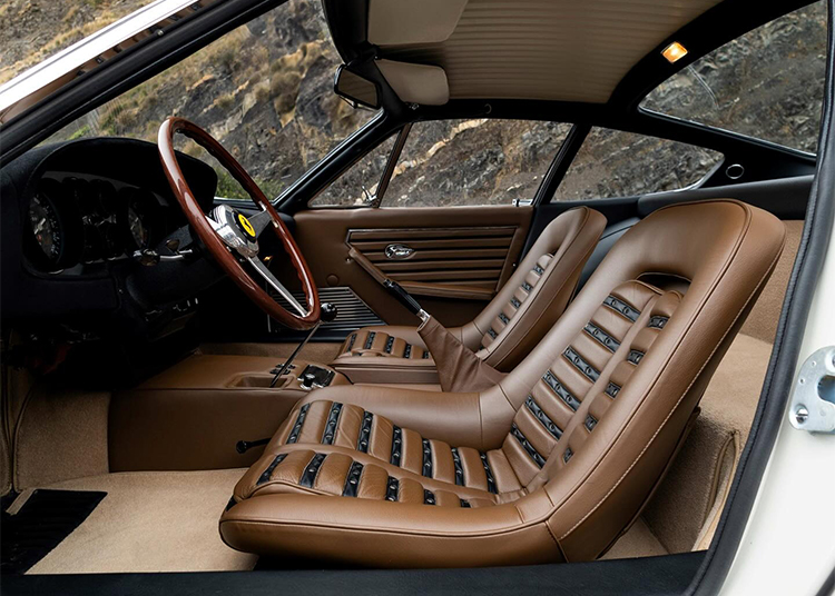 The image shows the interior of a vintage 365 GTB/4 Daytona with tan leather seats, black seat inserts, a wood-trimmed steering wheel, and manual gearshift, parked near a rocky hillside.