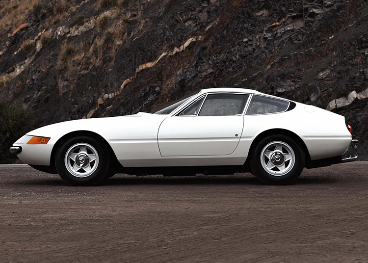 A classic white 365 GTB/4 Daytona with a sleek, low-profile design is parked on a paved road in front of a rocky hillside.