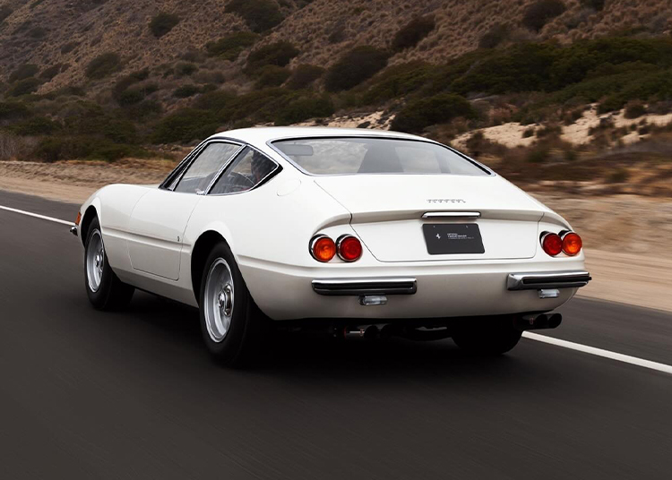 A classic white 365 GTB/4 Daytona sports car drives on a road beside a hillside covered with shrubs and sand.