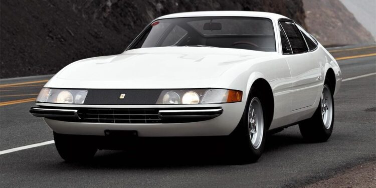 A white 365 GTB/4 Daytona classic sports car is parked on a winding mountain road with rocky cliffs in the background, exuding timeless elegance reminiscent of Ferrari Beverly Hills.