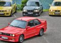 Four important BMWs—the E30 M3 in red, yellow, green, and yellow—are parked outdoors on asphalt with lush greenery in the background.
