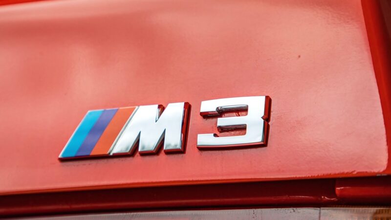 Close-up of a red BMW with an "M3" badge featuring blue, purple, and red stripes on the rear—an important symbol for E30 M3 enthusiasts.