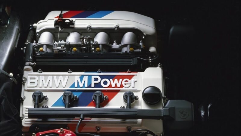Close-up view of an important BMW E30 M3 M Power engine with a white cover and blue, purple, and red stripes, displaying the BMW M Power logo on top.