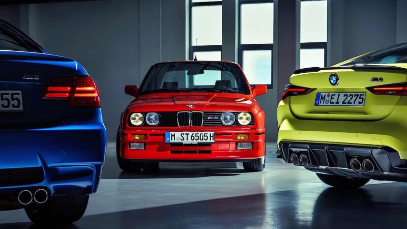 Three important BMW E30 M3 cars in blue, red, and yellow are parked indoors, showcasing front and rear views under artificial lighting.