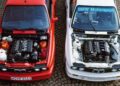 Two important BMW E30 M3 cars, one red and one white, are parked side by side with their hoods open, displaying detailed views of their engines on a cobblestone surface.