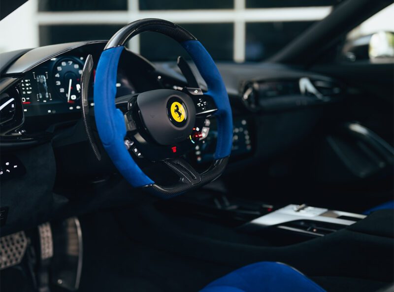 Close-up view of a modern Ferrari car interior featuring a black and blue steering wheel, digital dashboard, and metallic pedals in the stunning Nero DS Opaco finish, available at Miller Motorcars. Experience the power of 819HP 12Cilindri.