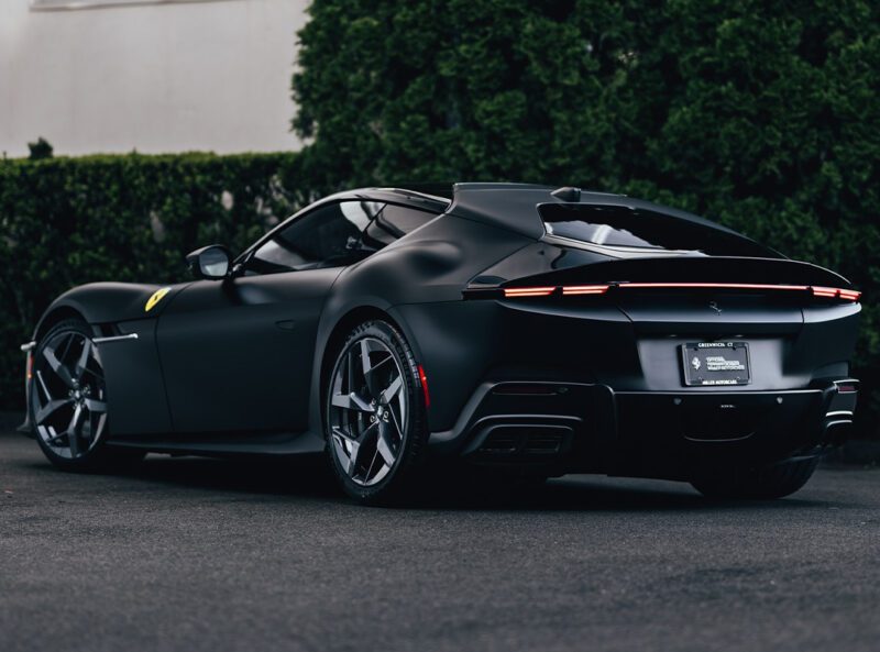 A matte black 12Cilindri luxury sports car with 819HP is parked on a dark pavement near a green hedge, viewed from the rear driver’s side—a striking presence worthy of Miller Motorcars.