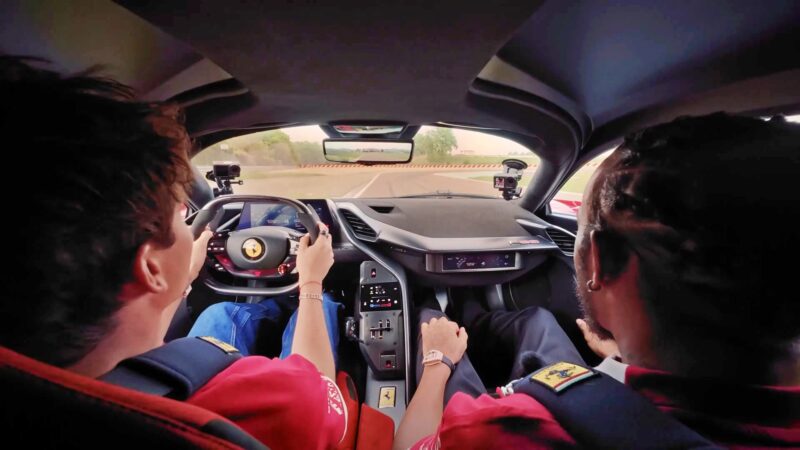 Hamilton and LeClerc, both wearing seatbelts, sit inside a Ferrari F80 as they track the Fiorano circuit, seen from the back seat with dashboard cameras capturing the drive.