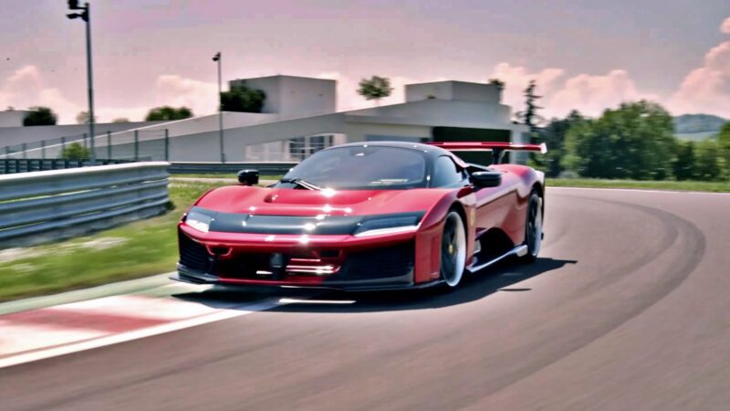 A red sports car drives at speed around a bend on the Fiorano racetrack, where Hamilton and LeClerc track the Ferrari F80, with modern buildings and greenery in the background.