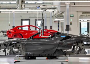 A black EV convertible chassis and a red Ferrari coupe chassis are positioned in a brightly lit automotive factory assembly area.