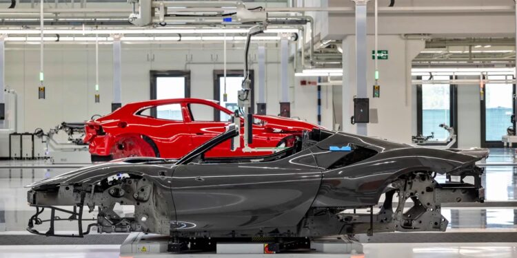 A black EV convertible chassis and a red Ferrari coupe chassis are positioned in a brightly lit automotive factory assembly area.