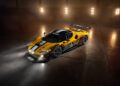 A yellow Ferrari 296 GT3 Evo with black and white stripes is parked indoors on a polished concrete floor, illuminated by overhead lights, reminiscent of a Ferrari unveiling at the 24 Hours of Spa.