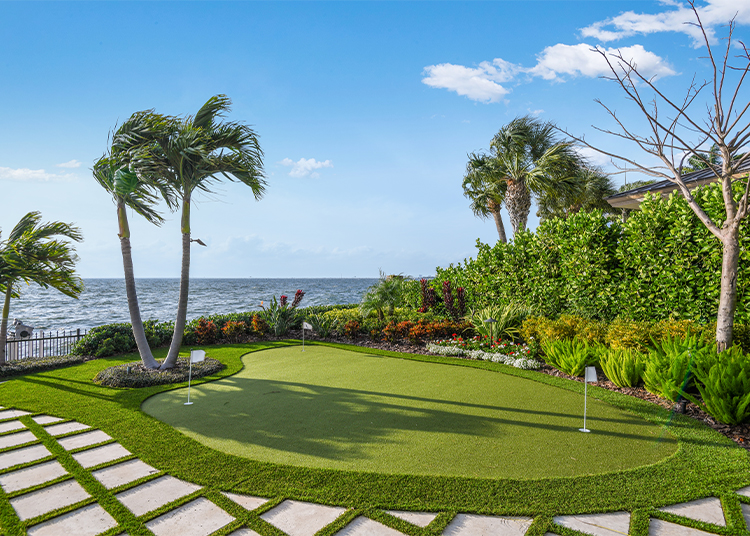 A backyard putting green with flags, surrounded by palm trees, plants, and a stone path, overlooks the ocean under a blue sky—perfect for those who shop for luxury home accessories and appreciate refined living.