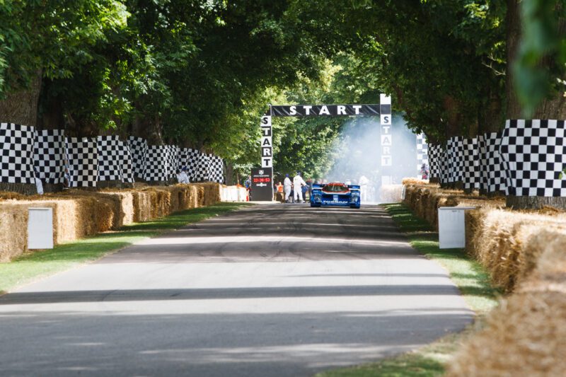A blue race car prepares to start at a tree-lined track with a "START" sign overhead and checkered flag banners—capturing the excitement of the Goodwood Festival of Speed, with TAG Heuer as Official Timing Partner.