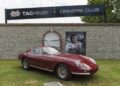 A classic red Ferrari sports car is parked on grass in front of a stone wall with TAG Heuer Drivers Club signage and a large racing driver poster at the Goodwood Festival of Speed, where TAG Heuer is the Official Timing Partner.