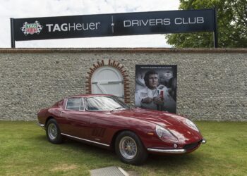 A classic red Ferrari sports car is parked on grass in front of a stone wall with TAG Heuer Drivers Club signage and a large racing driver poster at the Goodwood Festival of Speed, where TAG Heuer is the Official Timing Partner.