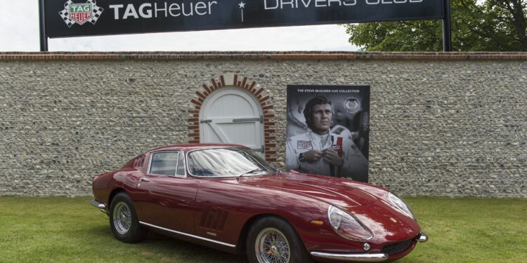 A classic red Ferrari sports car is parked on grass in front of a stone wall with TAG Heuer Drivers Club signage and a large racing driver poster at the Goodwood Festival of Speed, where TAG Heuer is the Official Timing Partner.