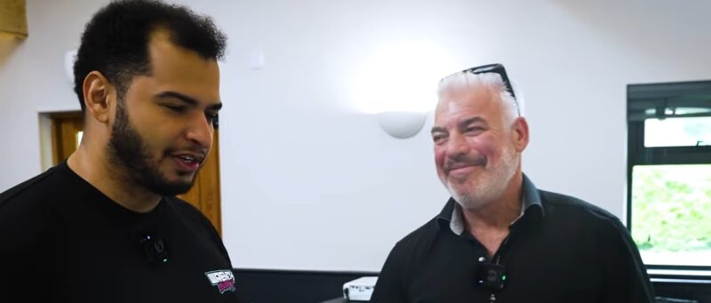 Two men stand indoors, engaged in conversation about Tavarish's flood-damaged McLaren P1 getting a wild reboot as a 1400 HP EVO variant by original designer Frank Stephenson. One smiles with sunglasses on his head; the other has a neutral expression.
