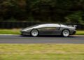 A sleek, silver Peralta S supercar drives at high speed on a racetrack, with blurred trees and tire barriers in the background, capturing the thrill of the Monterey Motorsports Festival.