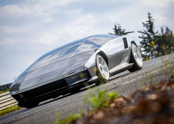 A futuristic, silver Peralta S supercar with a sleek, angular design is parked on a road with grass and trees in the background, evoking the innovation seen at events like the Monterey Motorsports Festival.