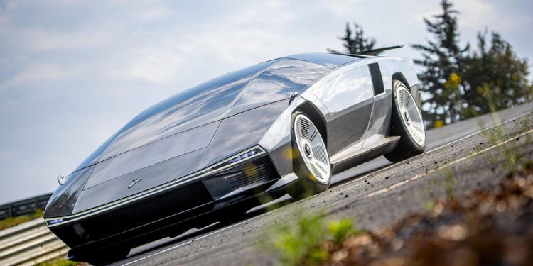 A futuristic, silver Peralta S supercar with a sleek, angular design is parked on a road with grass and trees in the background, evoking the innovation seen at events like the Monterey Motorsports Festival.