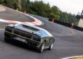 A silver Lamborghini supercar drives on a curving racetrack at the Monterey Motorsports Festival, surrounded by trees and safety barriers, with orange cones along the track edge.