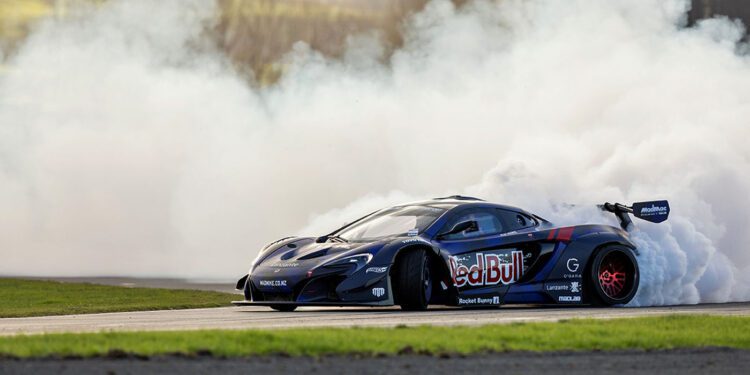 A black Red Bull Drift Car drifts on a track at the Monterey Motorsports Festival, producing a large cloud of white smoke from its rear tires.