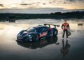A racing driver in a Red Bull suit stands on wet pavement next to a blue Red Bull race car at the Monterey Motorsports Festival, as cloudy weather sets the scene for thrilling moments reminiscent of a drift car showdown.