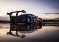 A McLaren P1 with Red Bull branding is parked on a wet surface at sunset, its striking silhouette reflecting on the ground.