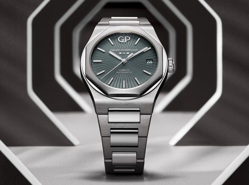 A silver Girard-Perregaux Laureato 50th Anniversary watch with a green textured dial is displayed upright against a geometric, hexagonal Infinite Grey background.