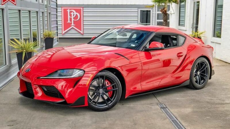 A red Toyota Supra sports car, one of the best sports cars under $100k, is parked on a concrete surface outside a modern building with large windows and potted plants. For sale now!.