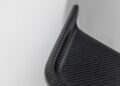 Close-up of a curved, black carbon fiber object with a textured, woven pattern—reminiscent of the finish seen on Gunther Werks Goes Modern: Meet GW9’s Carbon-Clad 992 Turbo S—set against a plain, light background.