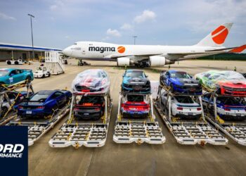 Multiple Ford sports cars, including the Mustang Dark Horse from Ford Performance, are secured on pallets in front of a Magma cargo airplane at an airport, ready for transport.