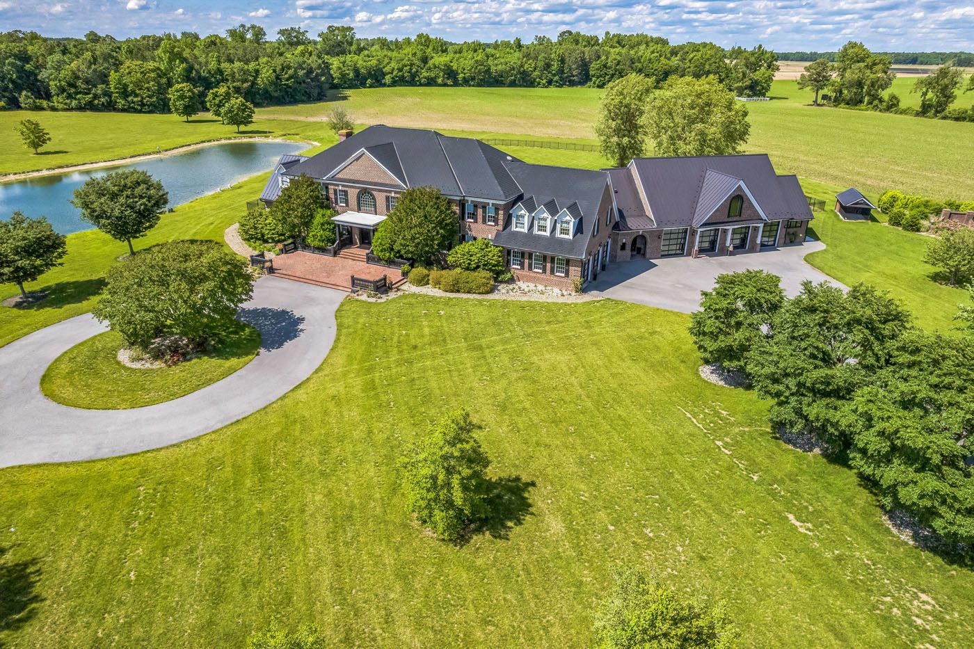 A large brick house with multiple gables and a circular driveway, situated on a spacious green lawn beside a pond, surrounded by trees and open fields—offering unique opportunities through U.S. Treasury Auctions.