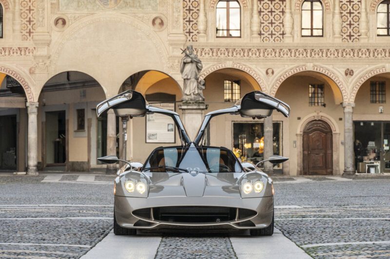 A silver Roadster with gullwing doors open is parked on a cobblestone plaza in front of a historic building with arched doorways and a statue, capturing the elegance and excitement of the MIMO Milano Monza Motor Show.