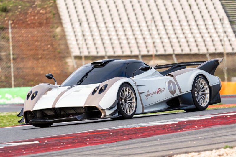 A white Pagani Huayra R Evo speeds along the racetrack at the Milano Monza Motor Show, with empty bleachers and a fence visible in the background.