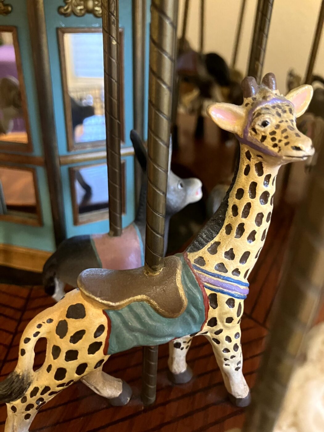 Close-up of a carousel figurine featuring a giraffe and a donkey, both with saddles, next to metal poles and a blue structure in the background.