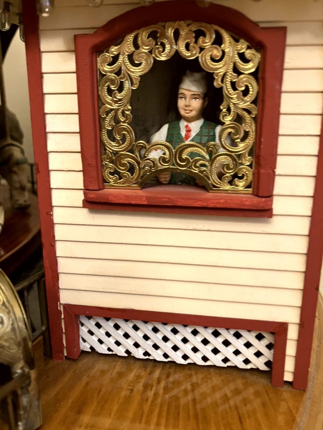 A figurine of a person in traditional attire is seen through a decorative gold-framed window on a model building with cream siding and red trim.