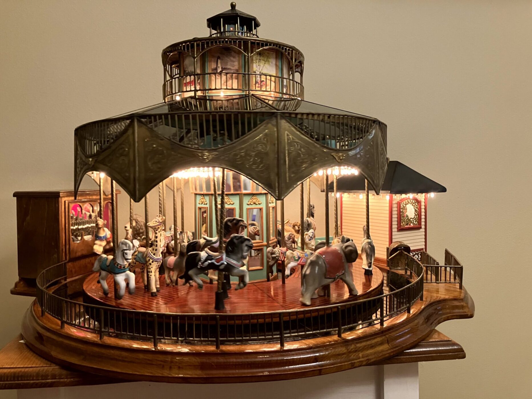 A detailed miniature carousel model featuring various animals, including horses, an elephant, and a zebra, displayed on a wooden base against a beige wall.