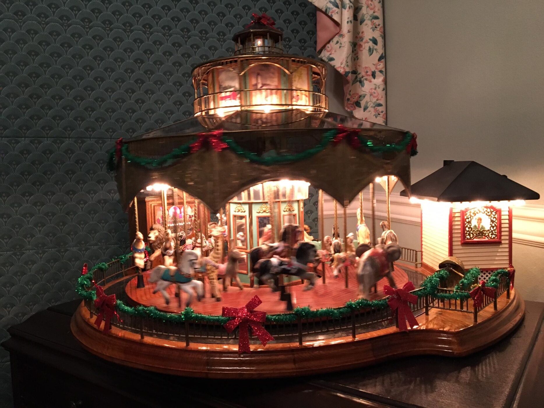 A miniature carousel decorated with red bows and green garlands sits on a dark wooden surface against patterned wallpaper.