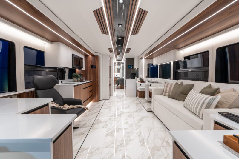 Luxury RV interior featuring marble-like flooring, a modern kitchen, lounge chair, sofa with pillows, and sleek wood-accented cabinetry under ambient lighting—perfect for a stylish escape in your Prevost motorcoach.
