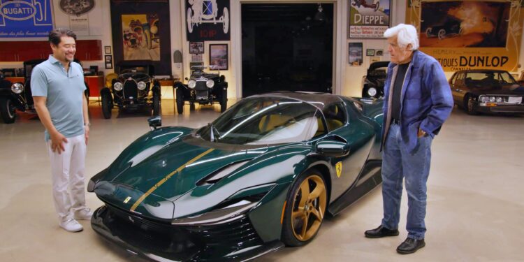 Two men stand beside a green Ferrari SP3 Daytona with gold accents in a garage filled with vintage car posters and classics, revealing just why Jay Leno is smitten by David Lee's stunning Ferrari masterpiece.
