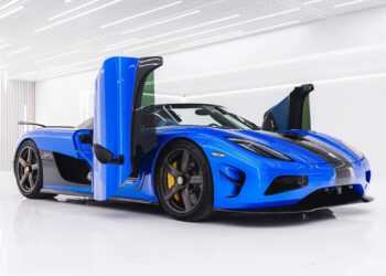 A Koenigsegg blue sports car with butterfly doors open, black details, and yellow brake calipers, parked indoors on a glossy white floor with bright overhead lighting—an iconic hypercar for sale.