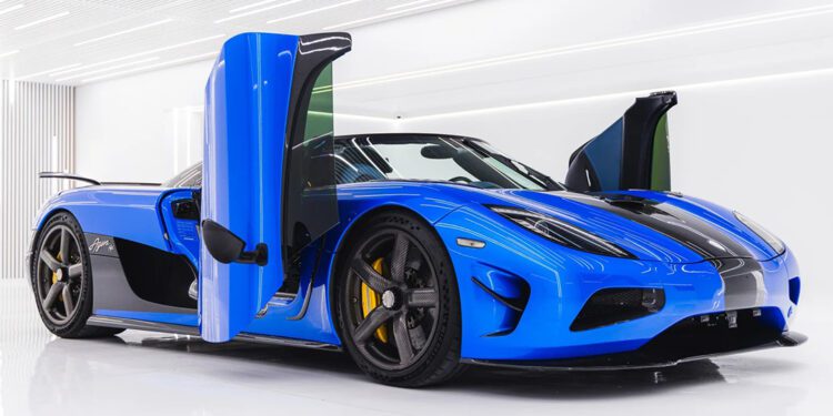 A Koenigsegg blue sports car with butterfly doors open, black details, and yellow brake calipers, parked indoors on a glossy white floor with bright overhead lighting—an iconic hypercar for sale.