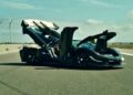 A black Farewell Jesko sports car with its butterfly doors and front trunk open is parked on a paved road near wind turbines under a blue sky, marking the arrival of the track-focused 1625 HP Koenigsegg Sadair's Spear.