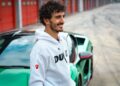 A man in a white Ducati hoodie stands near a green sports car—reminiscent of what two-time MotoGP champ Francesco Bagnaia makes of the Lamborghini Temerario—parked by red garage doors at a racetrack.