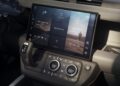 Close-up of the Land Rover Defender Refreshed: Here's What’s New For 2026 dashboard, featuring a large touchscreen with navigation, media controls, and climate control dials below. The gear selector and steering wheel are partially visible.